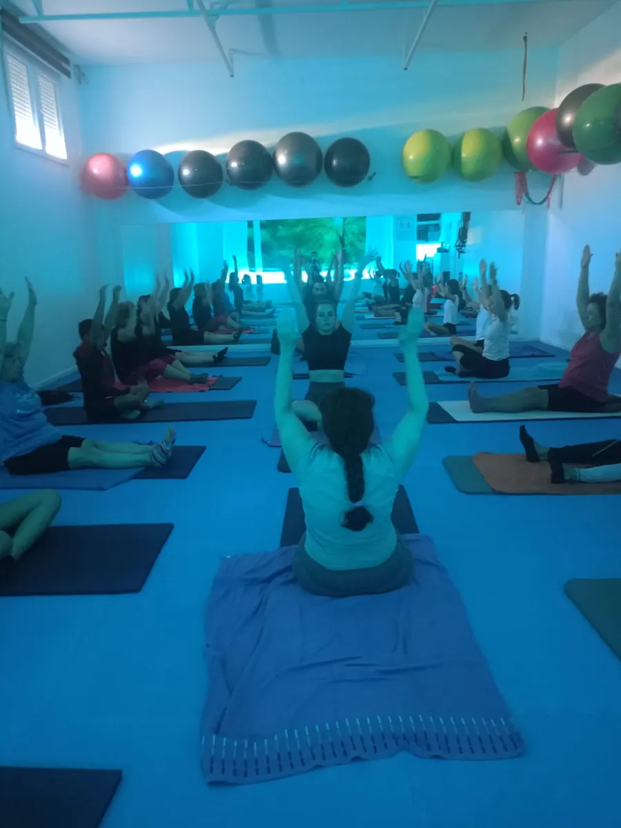 Clase de pilates en Loles Jiménez GYM & PILATES: un grupo numeroso sobre esterillas con los brazos en alto, con luz azul y fitballs colgados en la pared